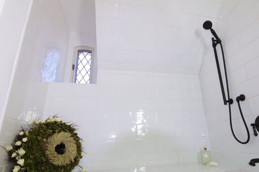 White tiled shower with black showerhead and hose, small window, wreath on the floor.