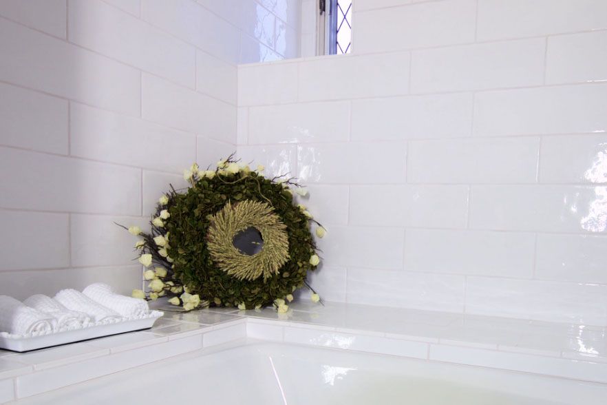 White tiled bathroom with a wreath leaning on the tub and rolled white towels on the ledge