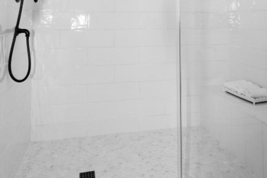 Minimal white tiled shower with glass partition, black handheld showerhead, and a folded towel on a shelf