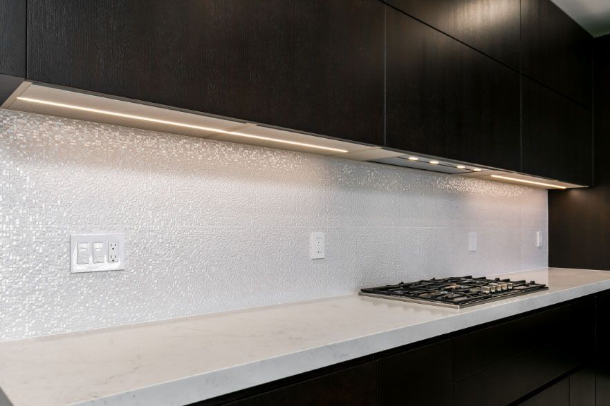 Modern kitchen countertop with dark cabinets, white backsplash, and gas cooktop under under-cabinet lighting