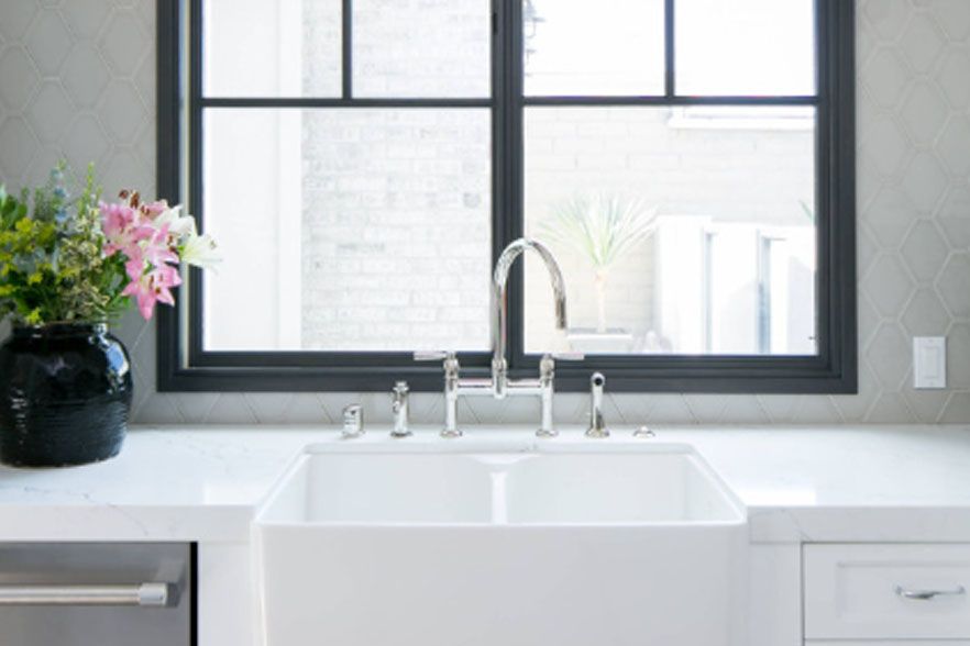 Modern kitchen sink beneath a large window, with white countertop and a vase of flowers on the left.
