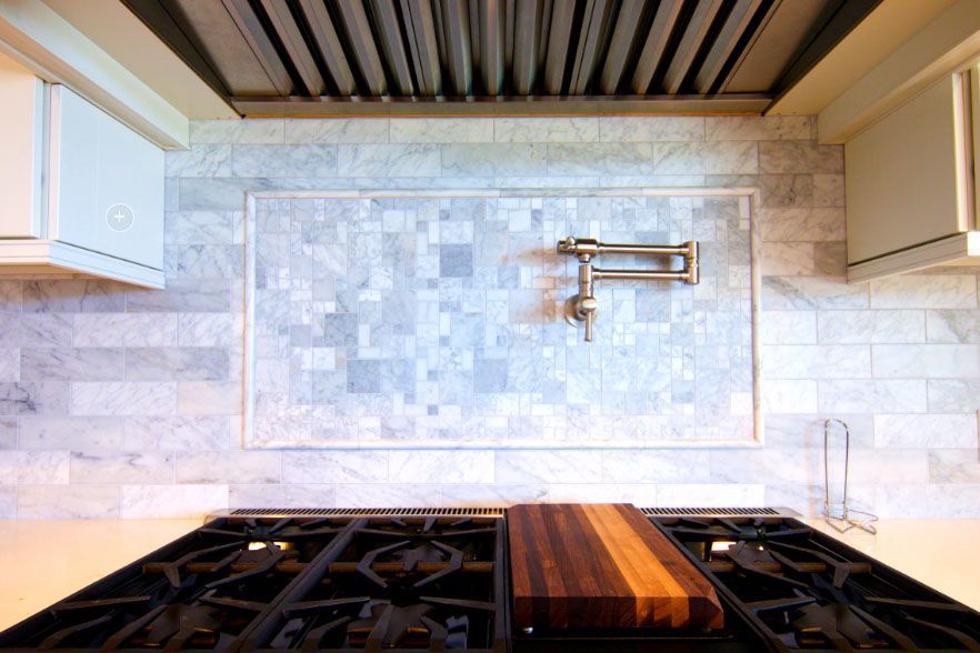 Modern kitchen with gas range, wood cutting board, and marble backsplash under a stainless hood