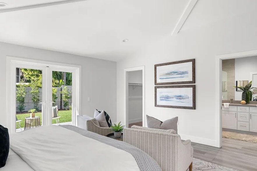 Bright bedroom with bed, lounge chair, framed art, and view of a green patio through French doors