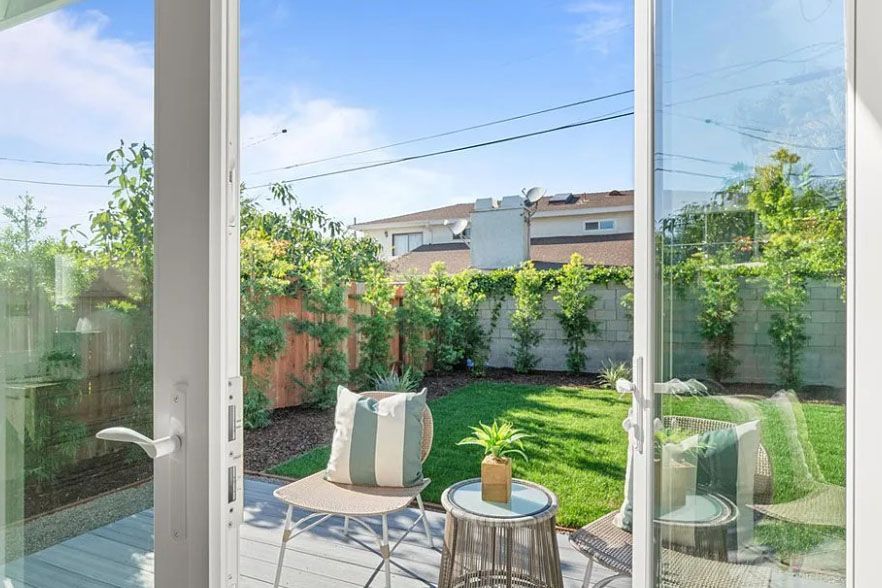 Sunny patio and green backyard seen through open glass doors, with outdoor chairs and small table.