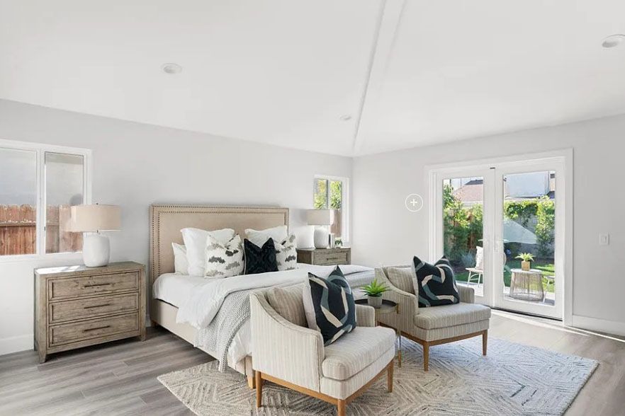 Bright bedroom with bed, upholstered chairs, wood dresser, and French doors to a sunlit patio.