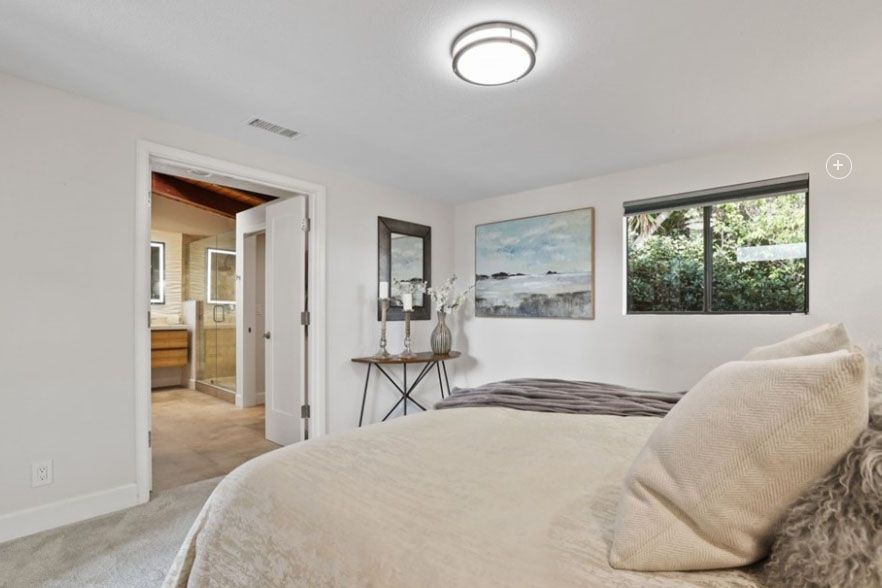 Bright bedroom with beige bedding, wall art, a small desk, and a view into a hallway and kitchen.