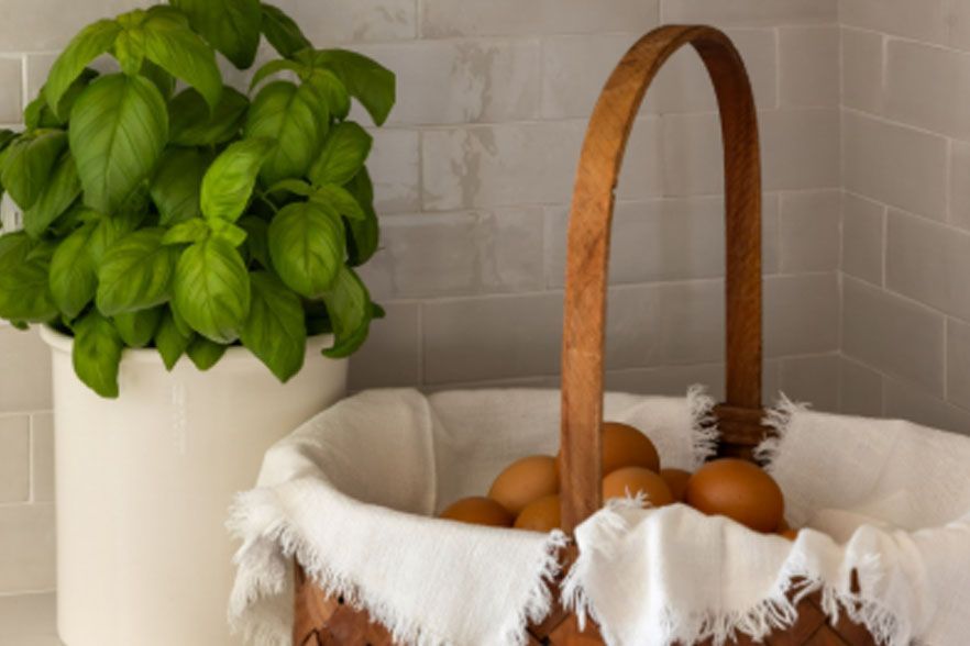 Basil plant in a white pot beside a basket of eggs on a tiled kitchen counter