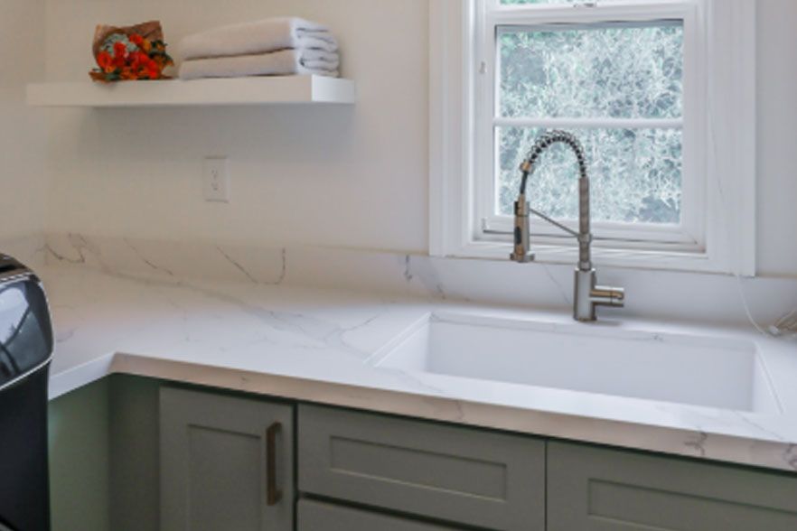 Bright kitchen sink under a window, with white counters, green cabinets, and a wall shelf with decor.