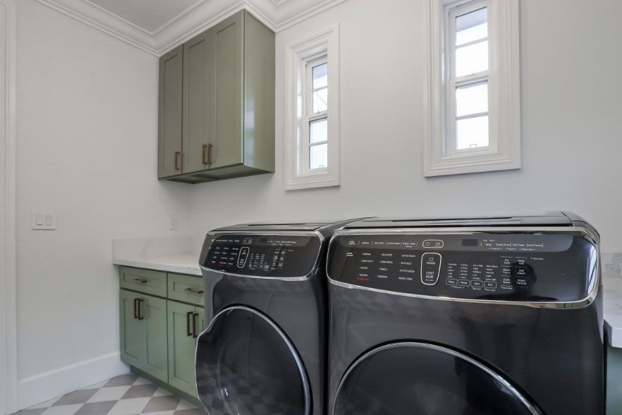 Bright laundry room with green cabinets, white walls, and black washer and dryer under two windows