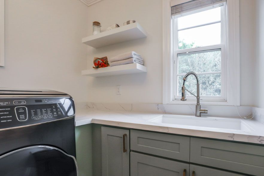 Laundry room with a black washer, white shelves, a window, and a gray sink cabinet under a faucet.