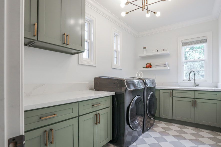 Bright laundry room with sage cabinets, white countertops, gray washer and dryer, and checkered floor.