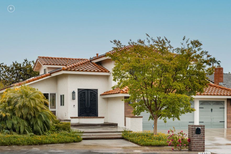 White stucco suburban house with red tile roof, front porch, garage, and a tree-lined yard