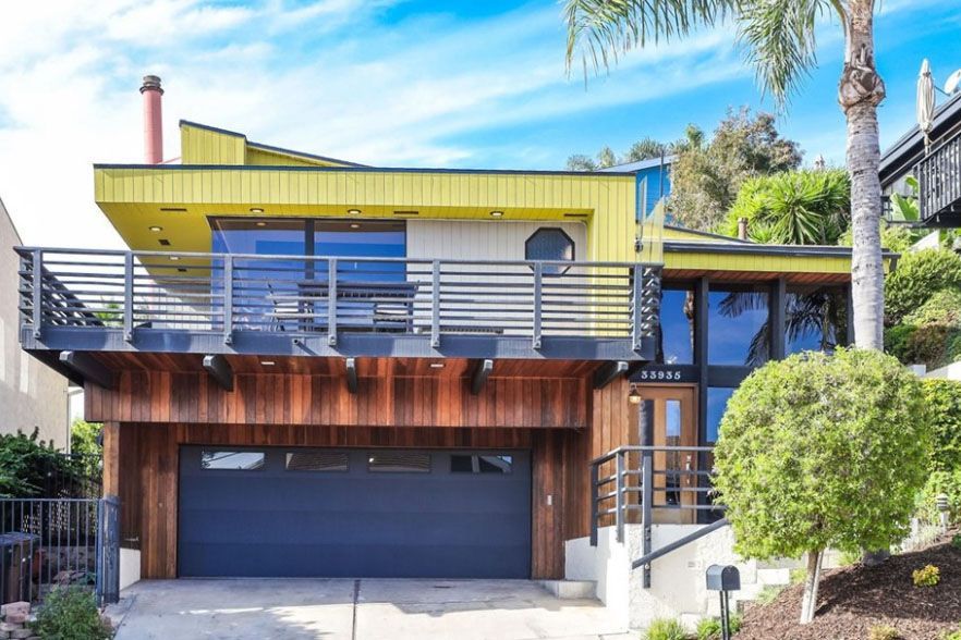 Modern two-story house with yellow siding, blue balcony, and dark garage under palm trees
