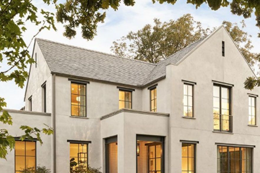 Modern white two-story house with large windows and a gray roof, framed by trees at dusk