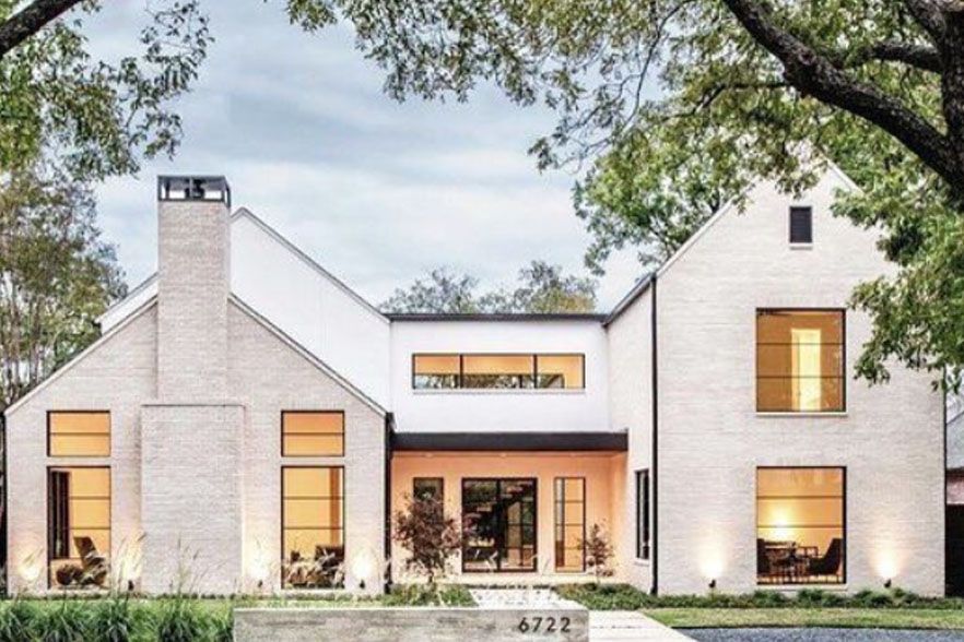 Modern white two-story house with warm interior lights and large front windows, framed by trees at dusk