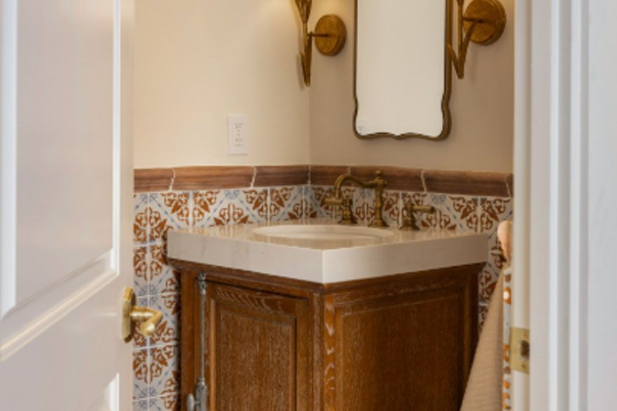 Small bathroom corner vanity with wood cabinet, white sink, mirror, and gold wall sconces