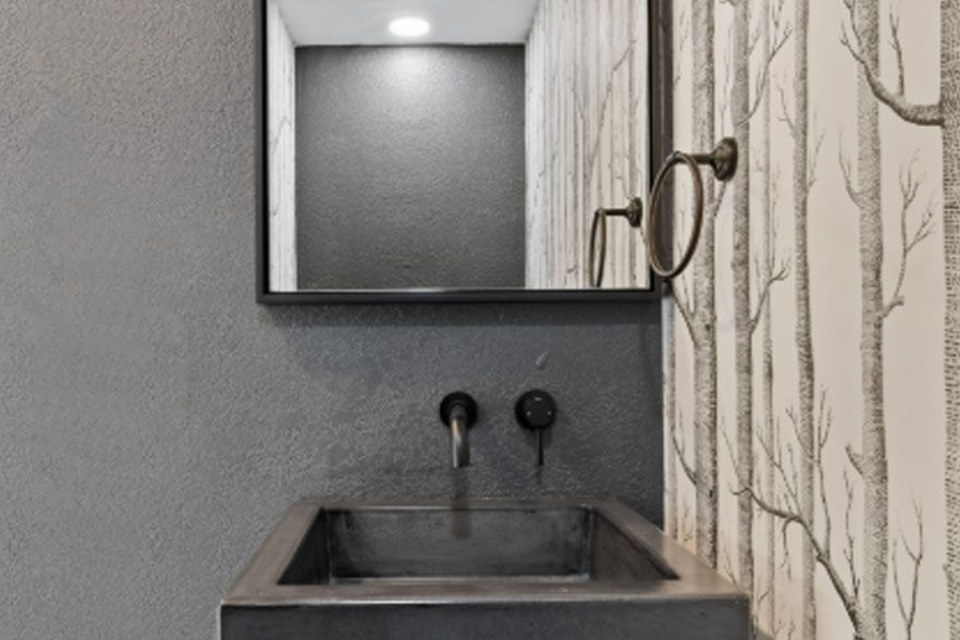 Small gray bathroom sink with mirror, wall sconce, and patterned wallpaper beside it.