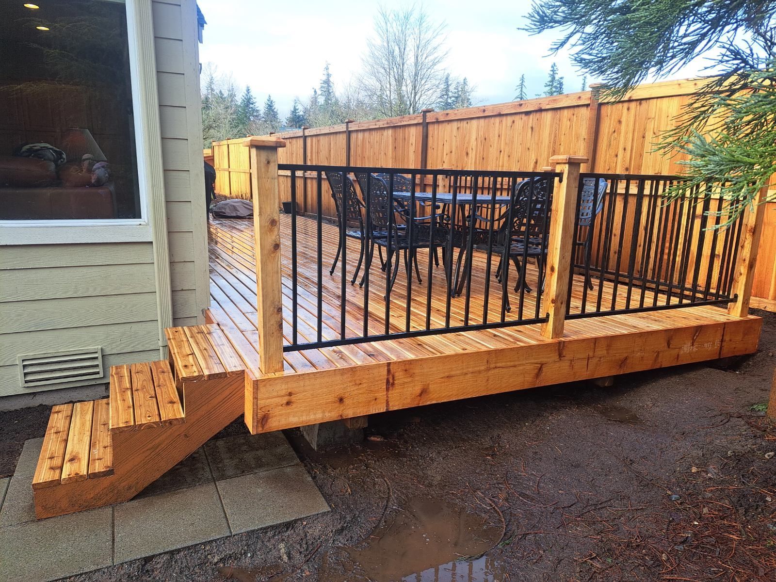 Wooden backyard deck with black railing and stairs beside a house, set on a gravel patio.