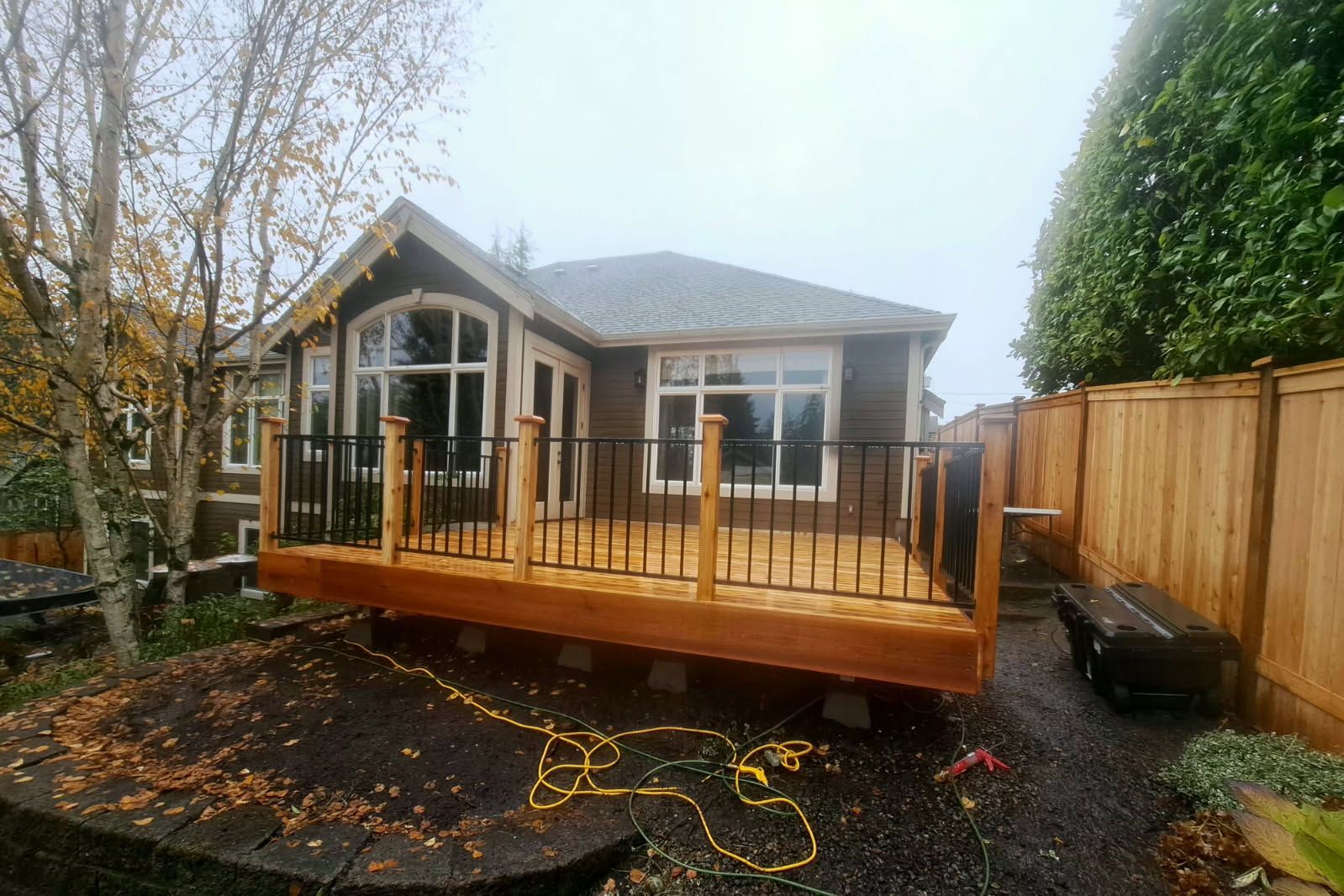 Backyard deck under construction beside a house, with wood railings and steps, surrounded by fencing and trees