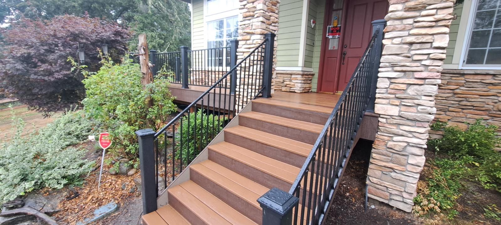 Front porch with stone columns, brown steps, black railings, and a red front door by landscaped shrubs