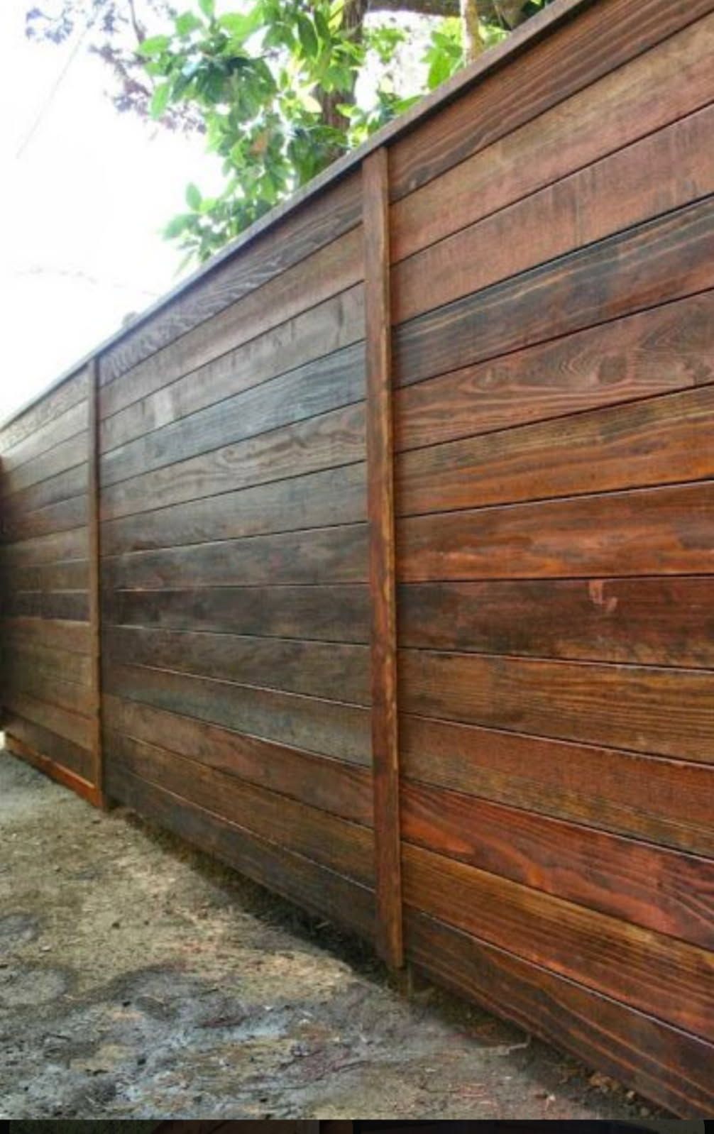 Wooden privacy fence with horizontal planks along a dirt path, under leafy trees