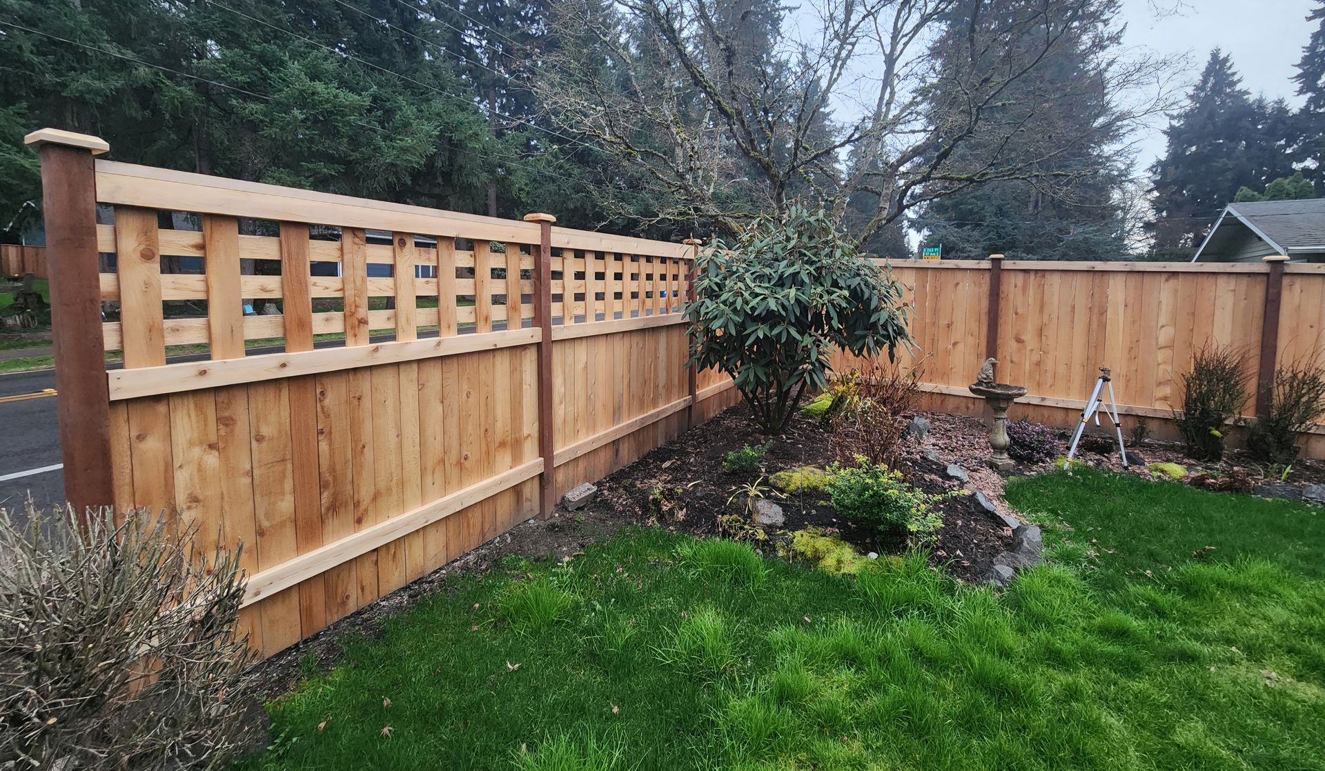 Wooden backyard fence with lattice top beside a green lawn and landscaped garden bed under overcast skies