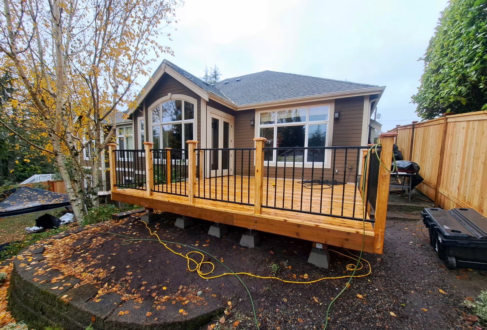 Small modern house with a wooden front deck and railing in a backyard, covered in autumn leaves.