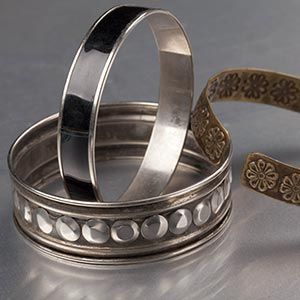 Three silver metal bangles, one with circular cutouts and one with a floral pattern, stacked on a gray surface