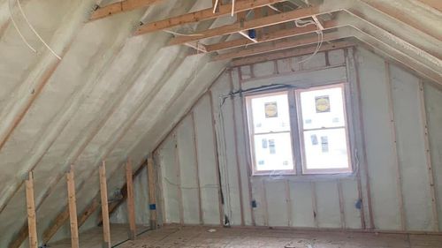 Unfinished attic with sloped insulated walls, exposed framing, and a small window at the far end