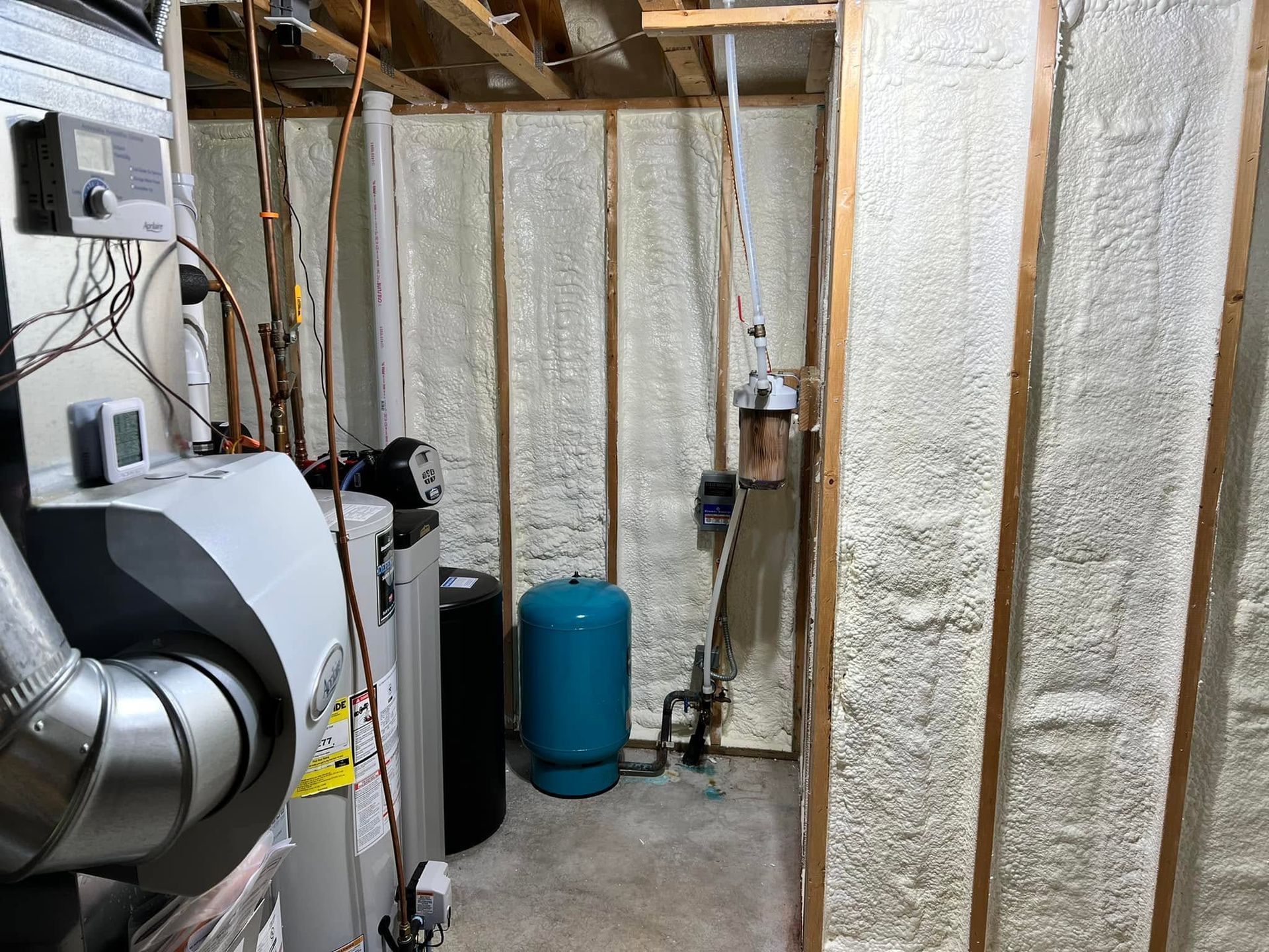 Basement mechanical room with exposed insulation, pipes, furnace equipment, and a blue water tank.