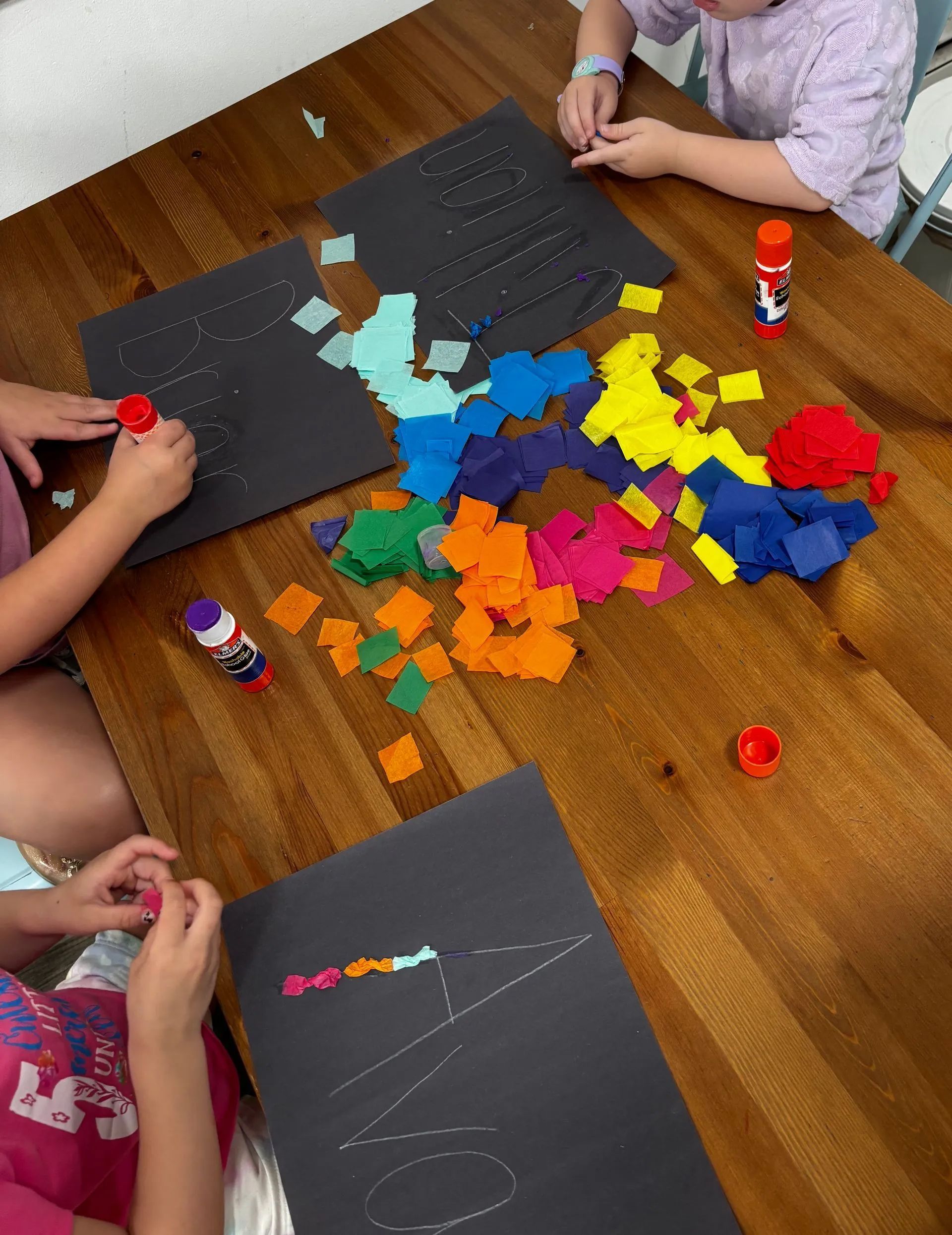 Children at a table create art with colorful squares on black paper using glue sticks.