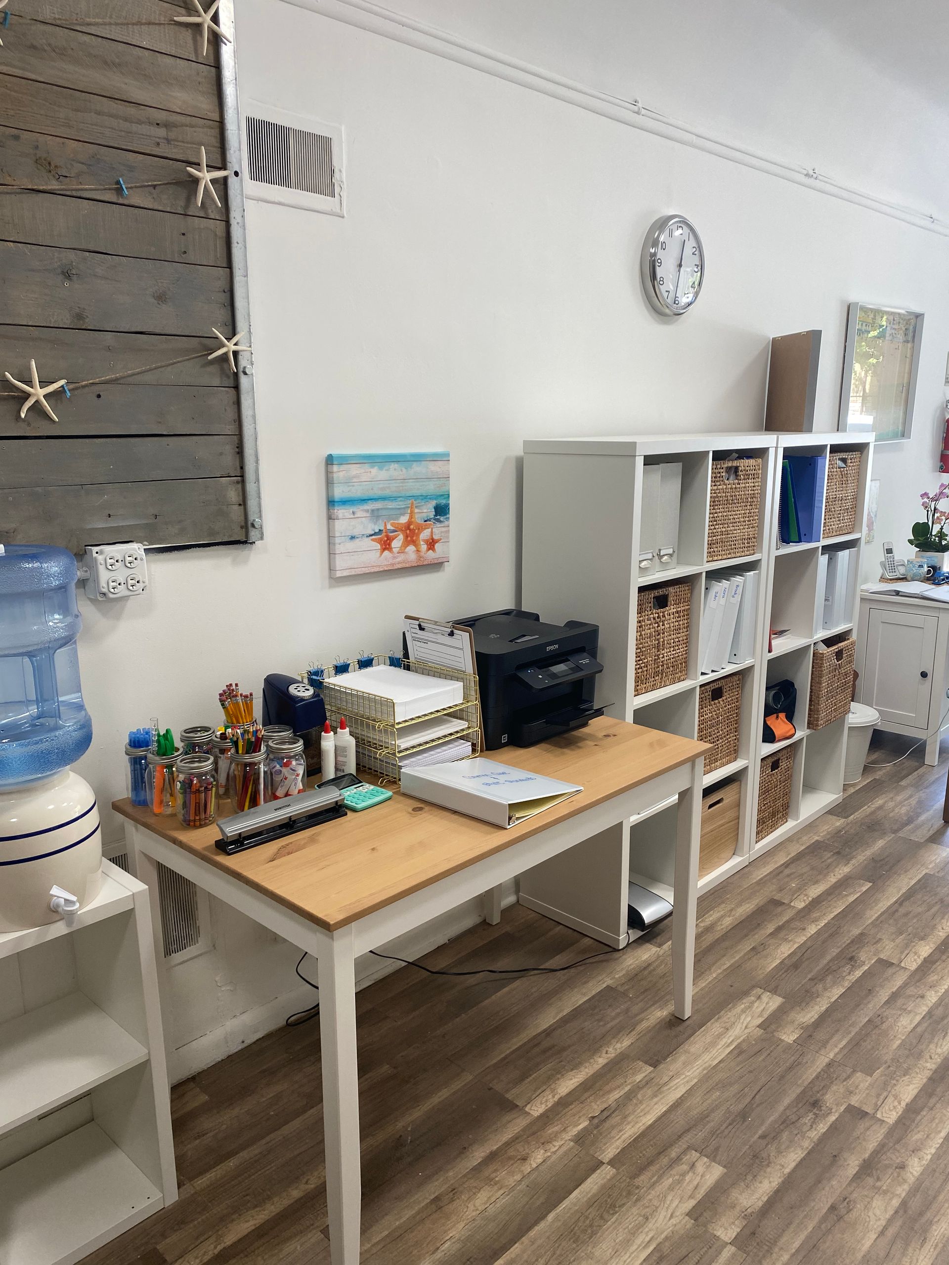 Office workspace with desk, printer, shelves, water cooler, and decorative items.