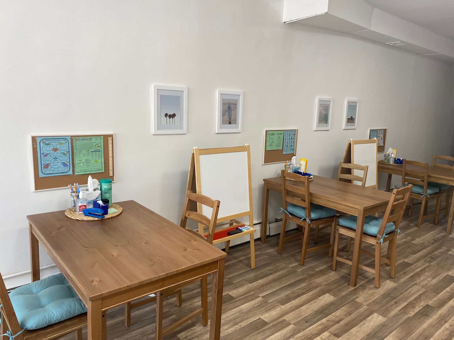 A room with wooden tables, chairs, and art supplies. Whiteboard and artwork on the walls.