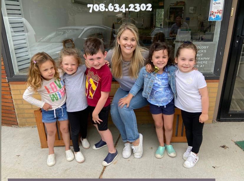 A woman sits with five children on a bench in front of a storefront. They are smiling.
