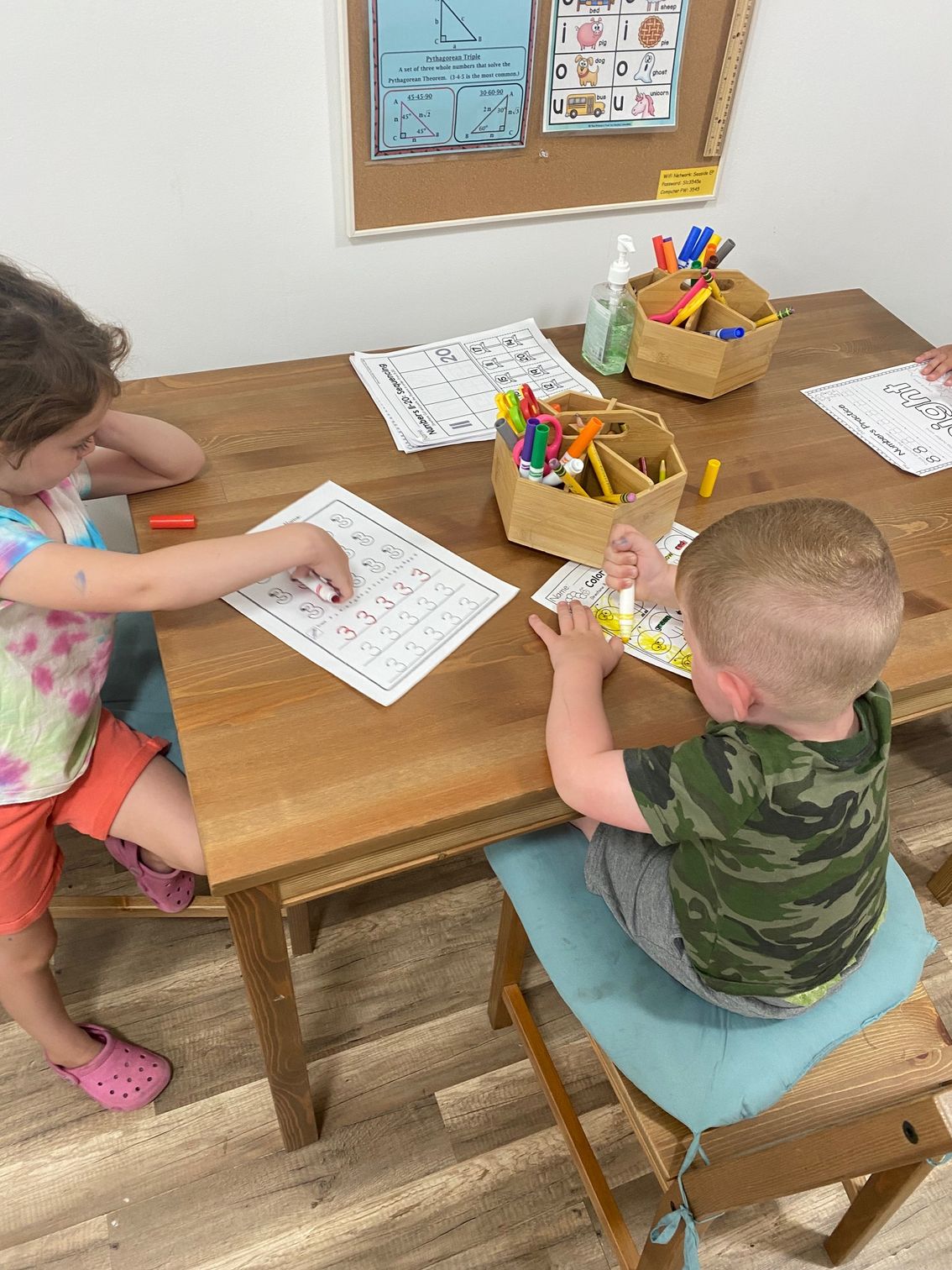 Two children at a wooden table, working on papers. Markers and pencils in a caddy are nearby.