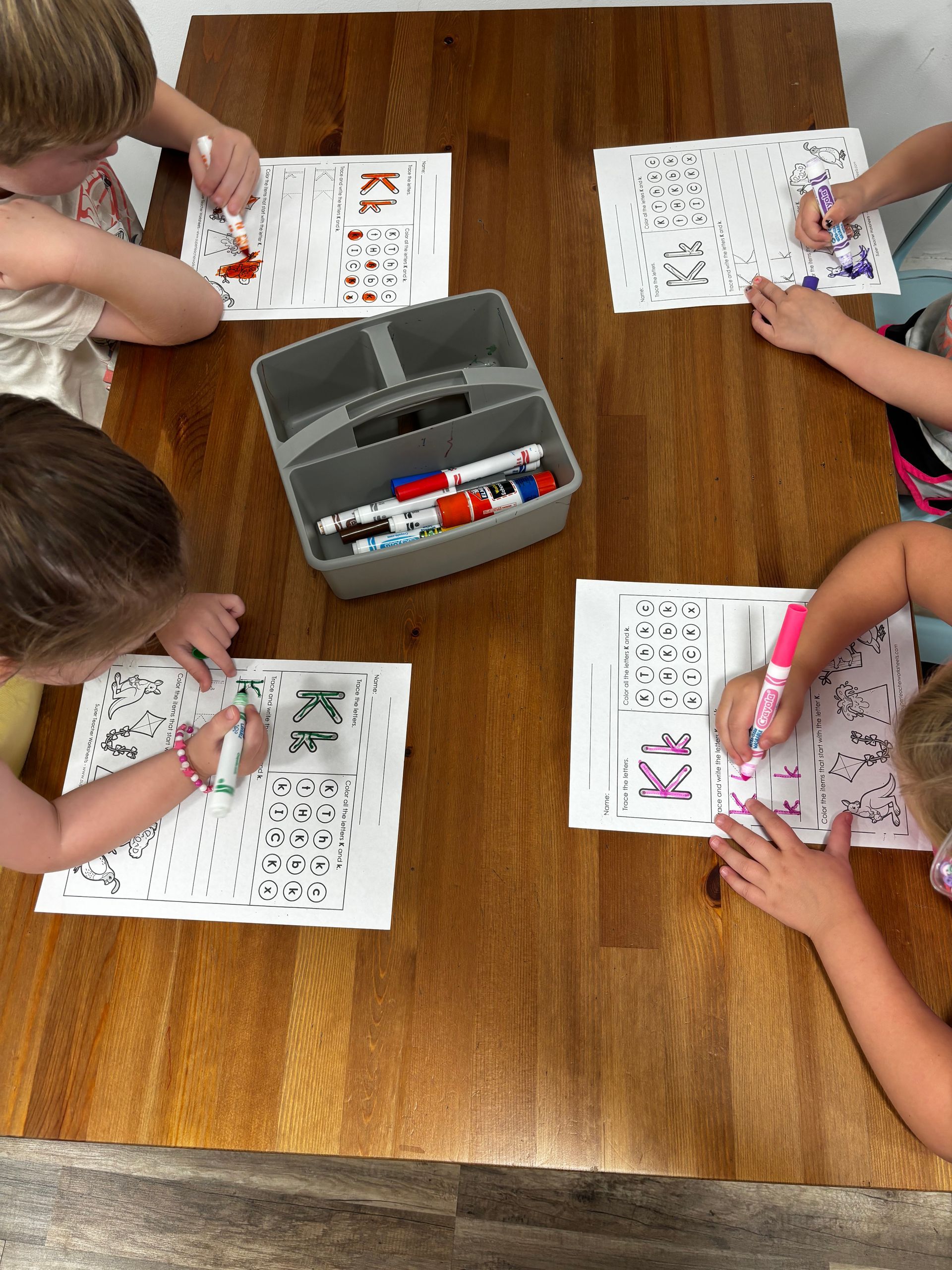 Children coloring on papers at a wooden table, using markers from a container.
