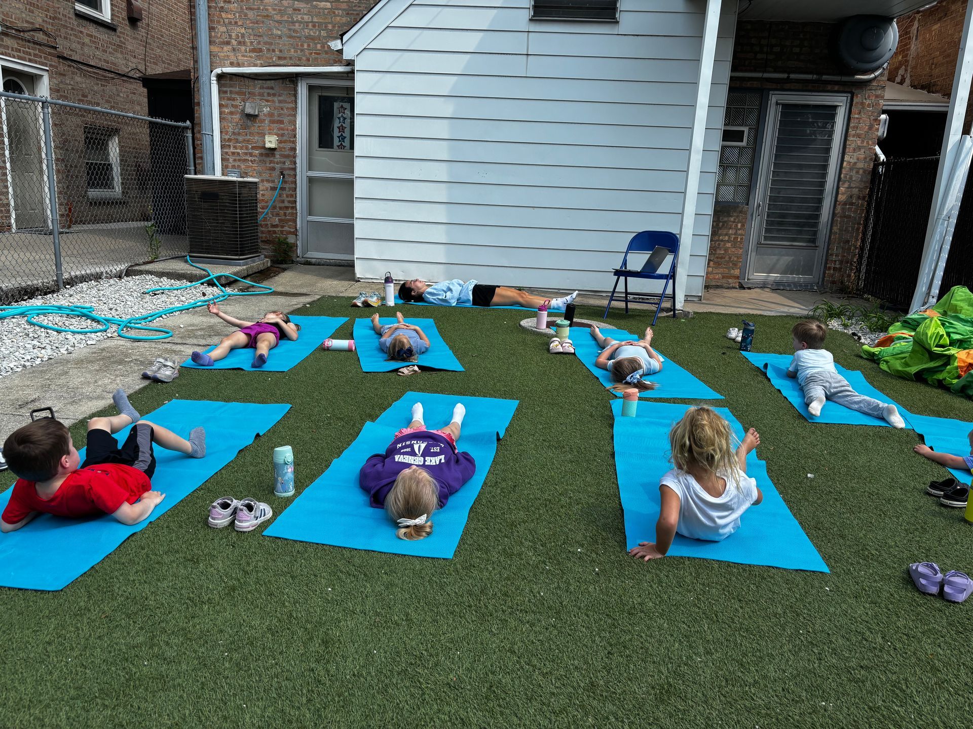 Children doing yoga on blue mats outdoors, white building in the background.