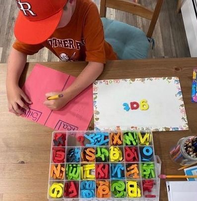 Boy in orange hat and shirt writing on pink paper at table with alphabet magnets.