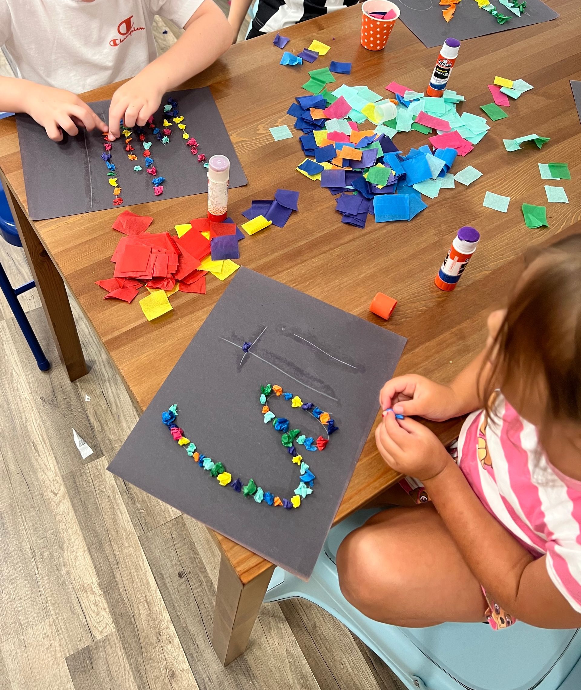 Children making mosaic art with colorful paper pieces on a wooden table.