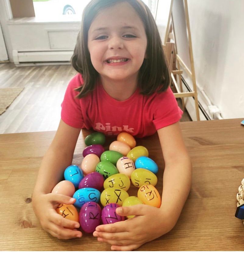Girl smiling, holding colorful Easter eggs with letters on a wooden table.