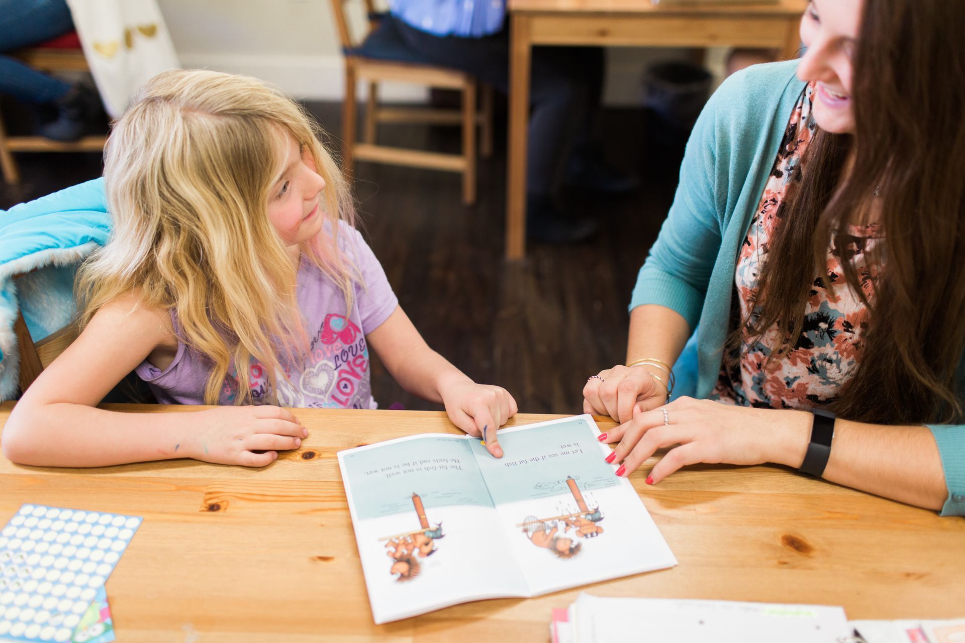 Girl and woman looking at a book together at a wooden table. The girl points at images.