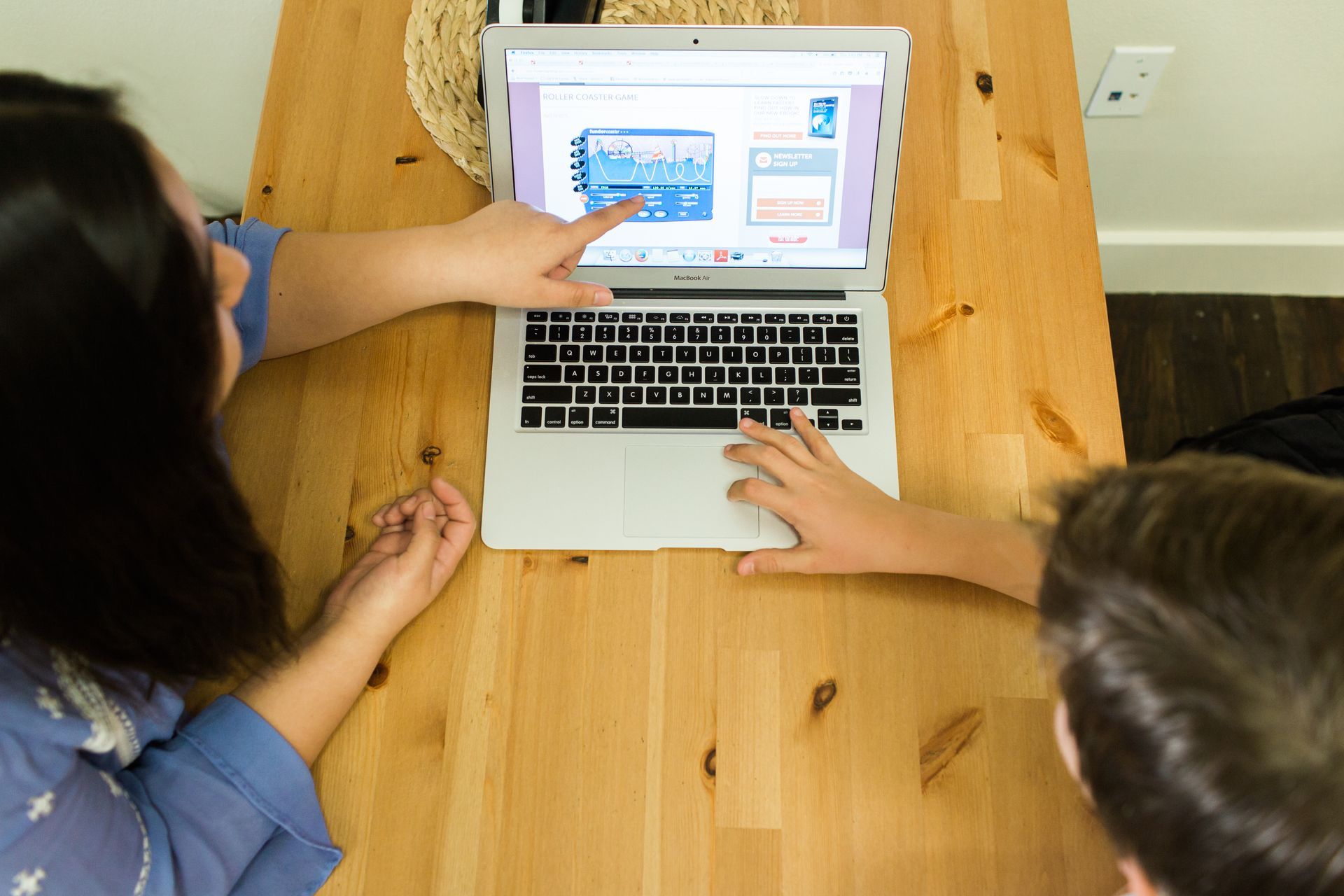 Person pointing at laptop screen while seated with another person at a wooden table.