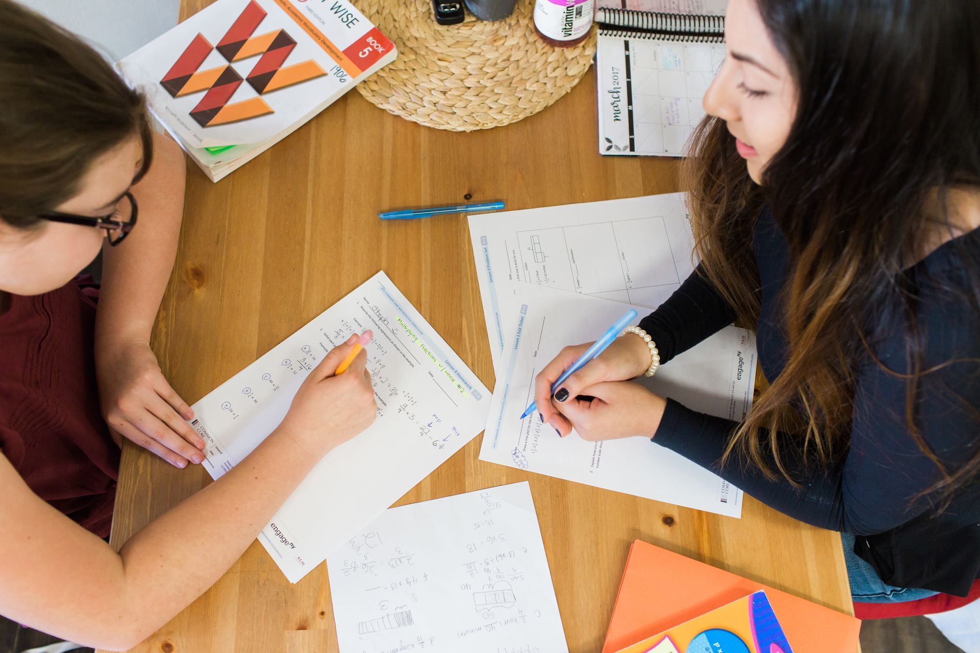 Two people at a wooden table work on papers, focused, with colorful books and supplies.