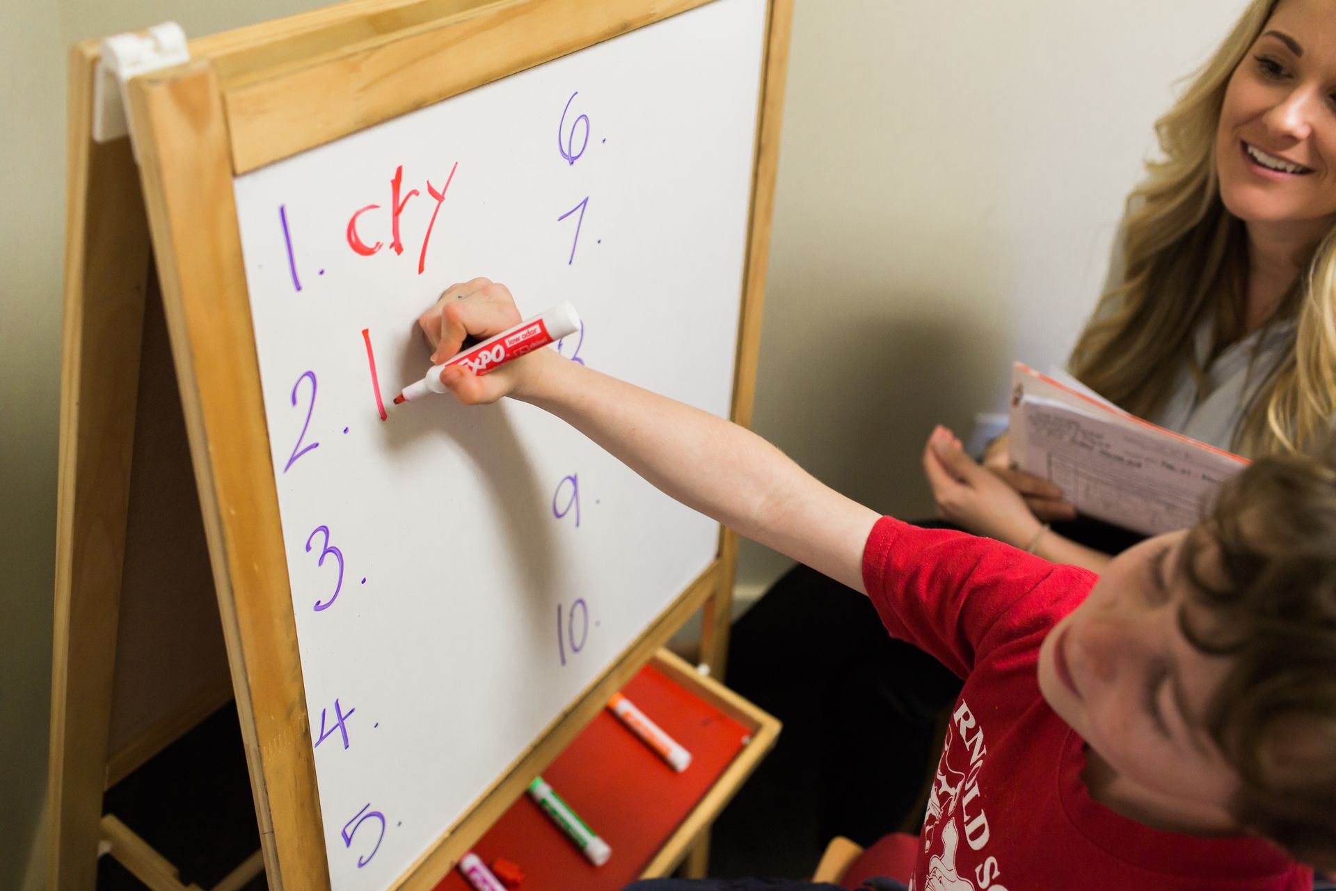 Child writing on a whiteboard with a red marker, assisted by a woman holding a notebook.