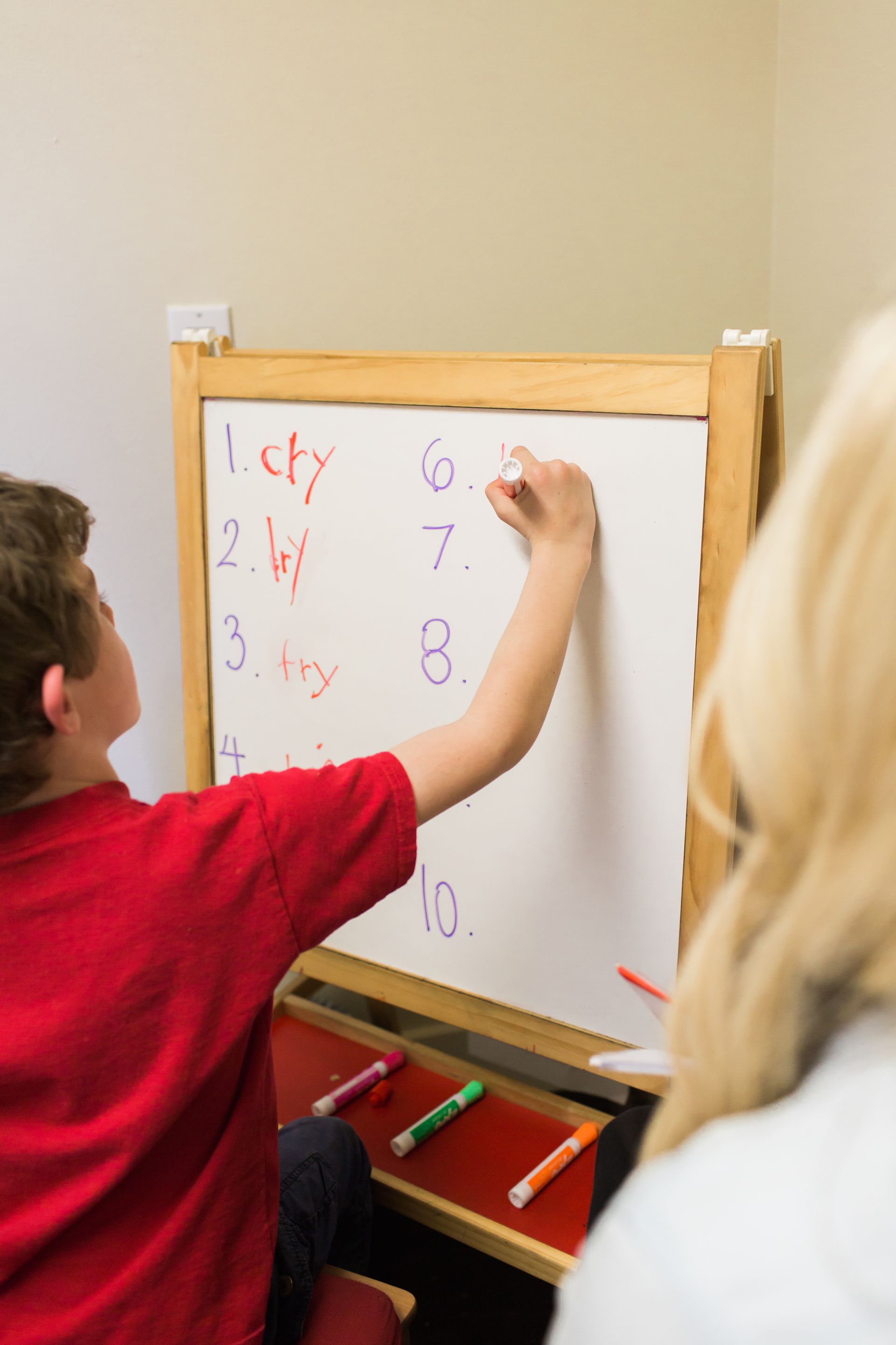 Boy in red shirt writing on a whiteboard with a numbered list, observed by a person.