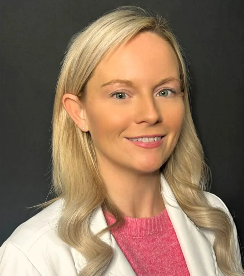 Portrait of a smiling person with blonde hair wearing a white coat and pink top on a dark background