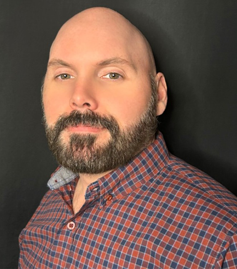 Portrait of a bearded man in a red-blue plaid shirt against a dark background