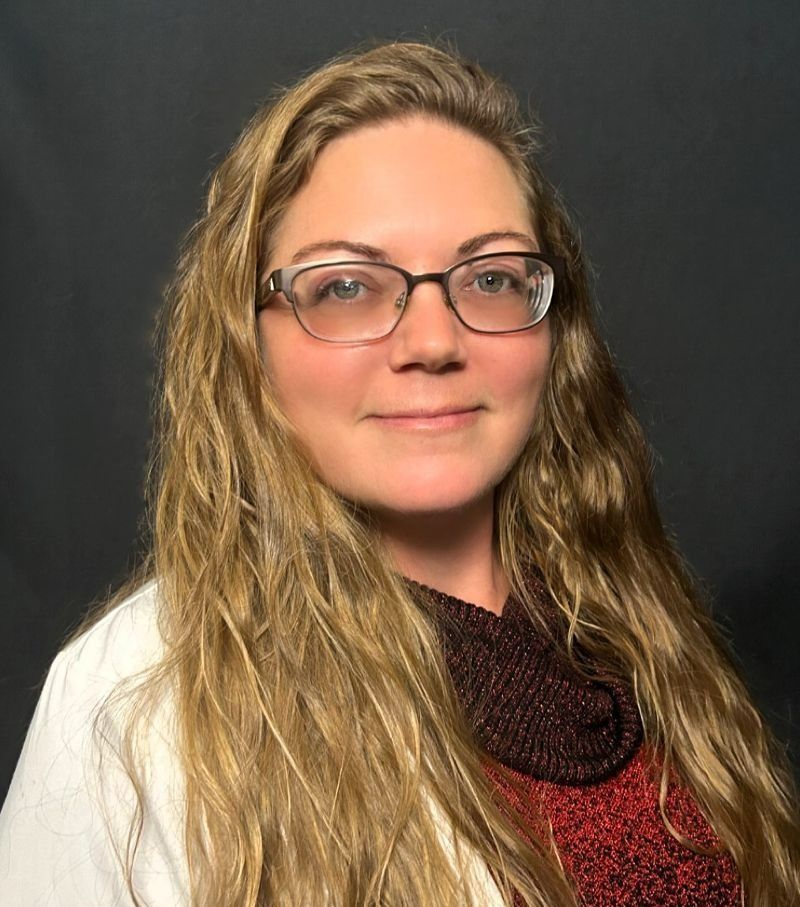 Portrait of a person with glasses and long wavy hair, wearing a black-and-red top against a dark background