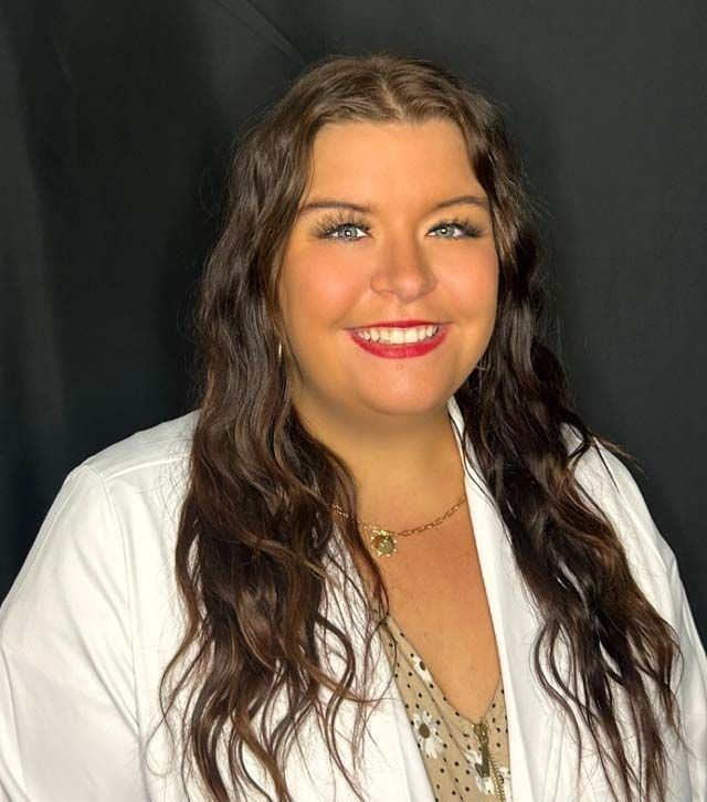Smiling woman with long wavy hair, wearing a white blazer and patterned top against a dark background