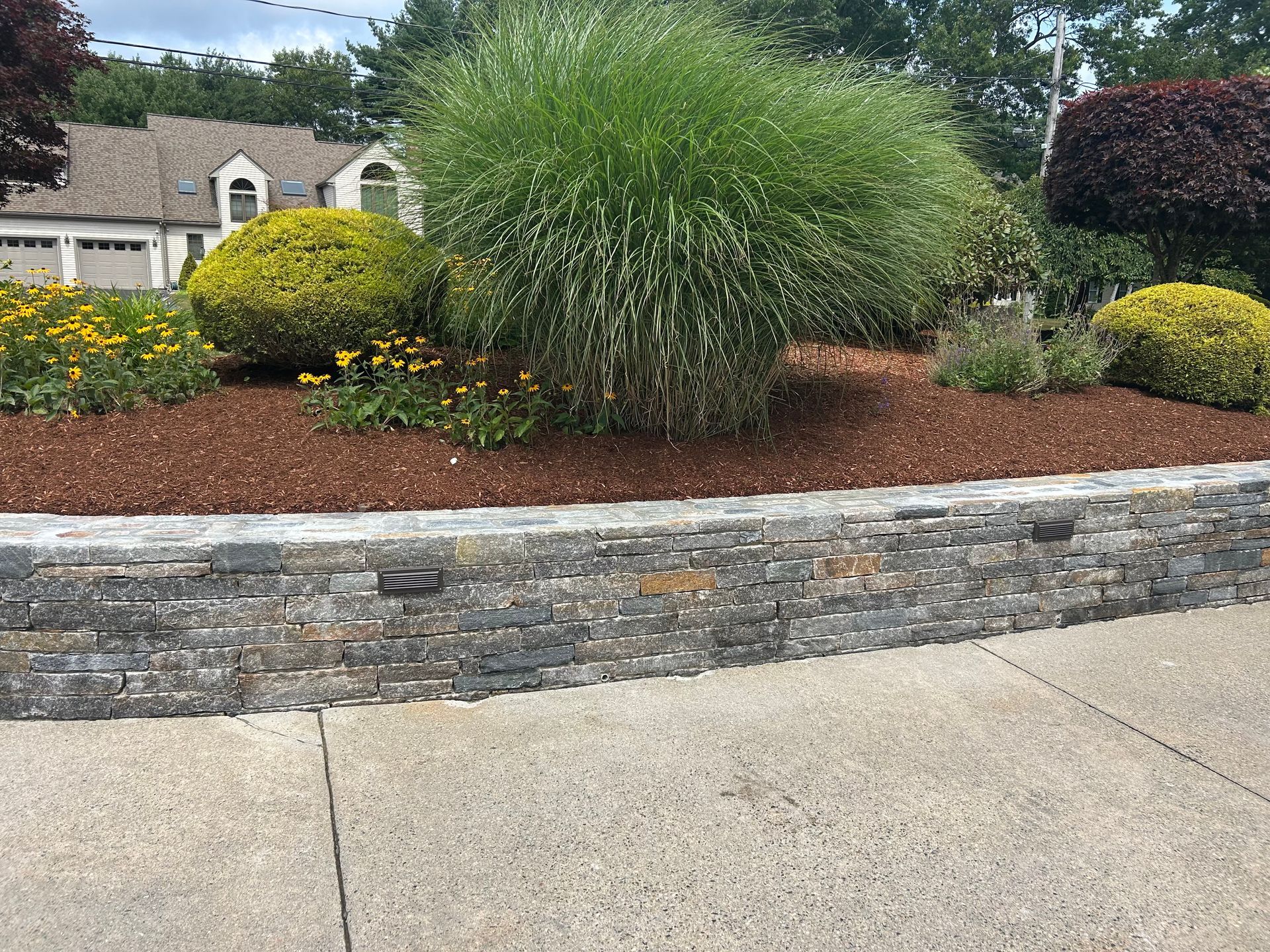 Landscaped garden bed with tall grasses, yellow flowers, and a stone retaining wall beside a sidewalk